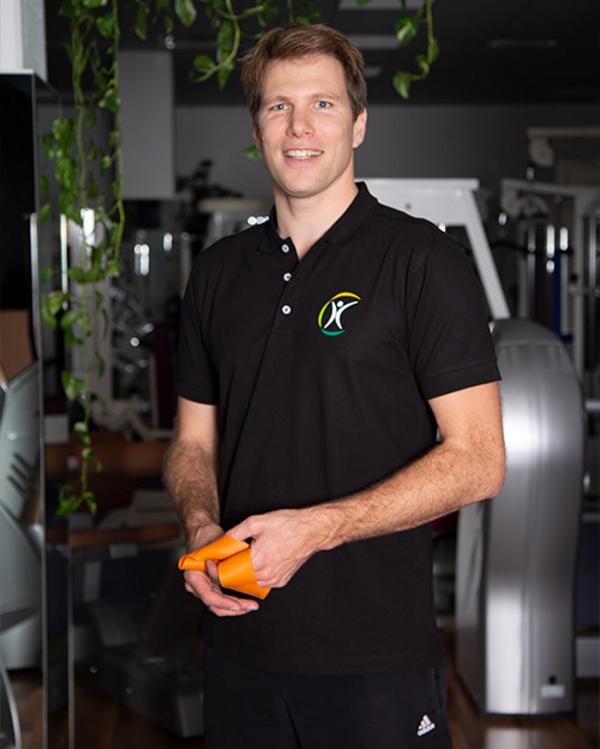 Trainer im Fitnessstudio hält ein orangenes Fitnessband und lächelt in die Kamera