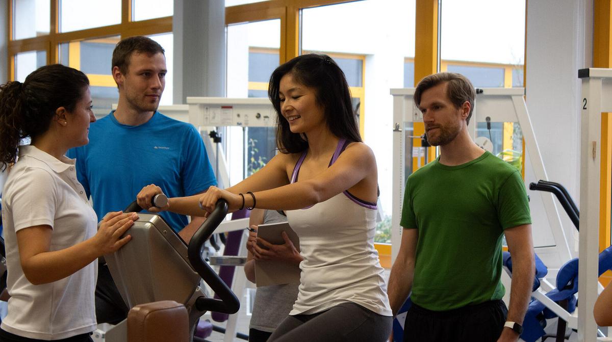 Frau trainiert auf einem Ergometer, drei Personen beobachten und beraten sie