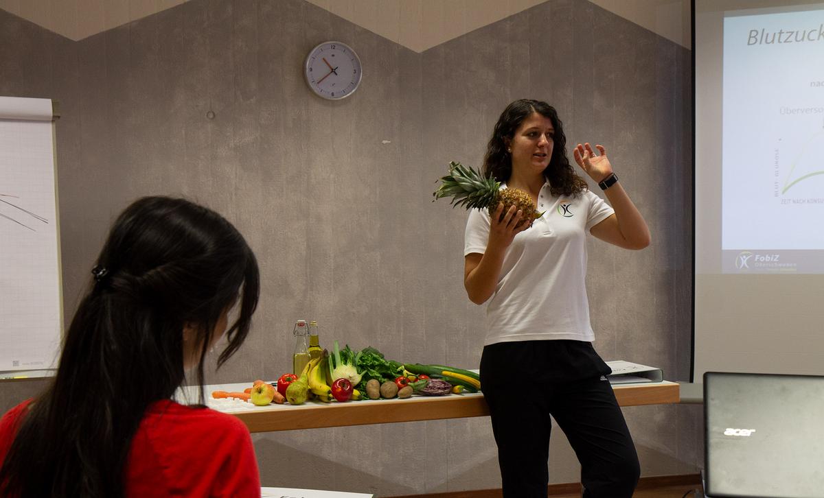 Dozentin hält eine Ananas hoch und spricht vor einer Auslage mit Obst und Gemüse