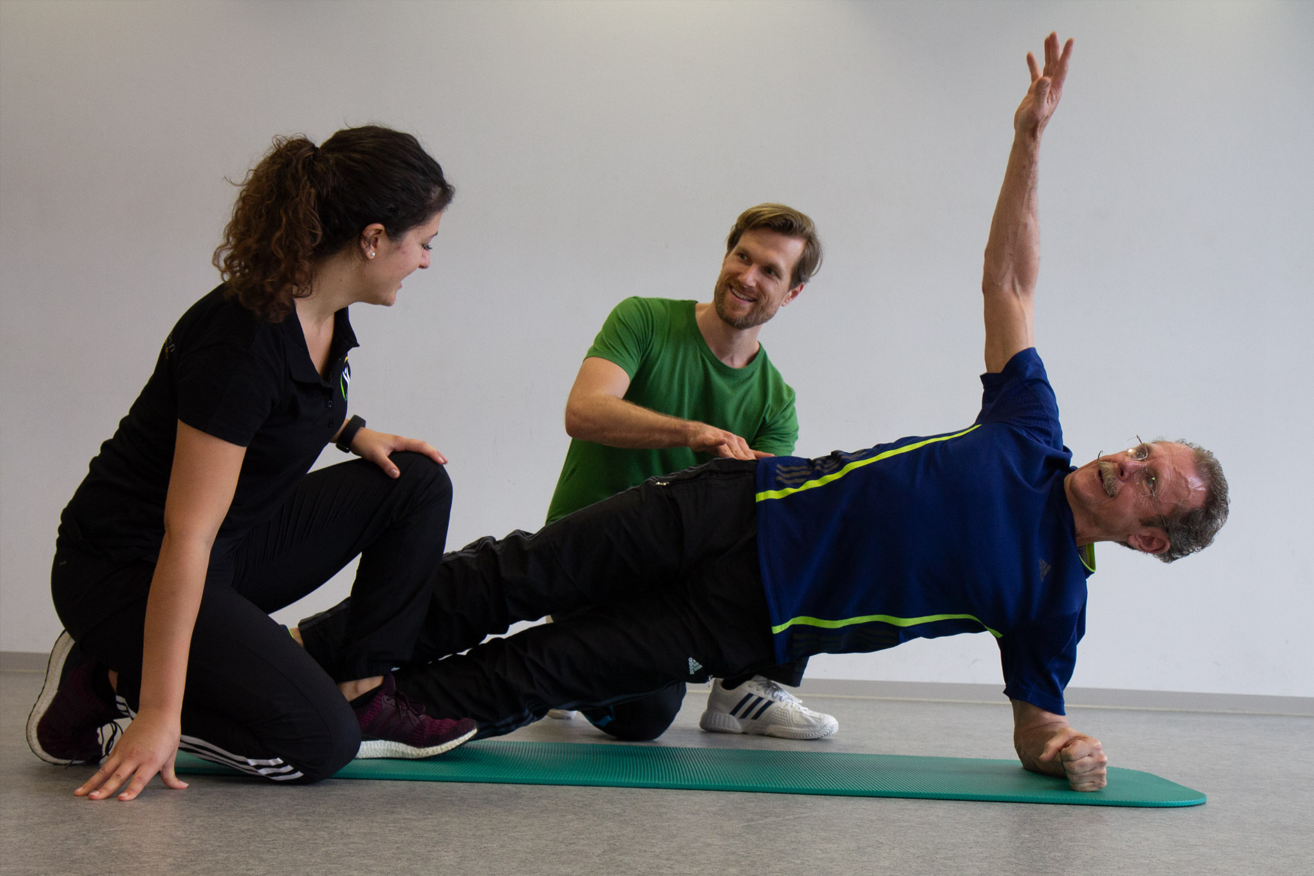 Älterer Mann macht seitliche Plank-Übung, Trainerin stützt, Trainer gibt Anweisungen