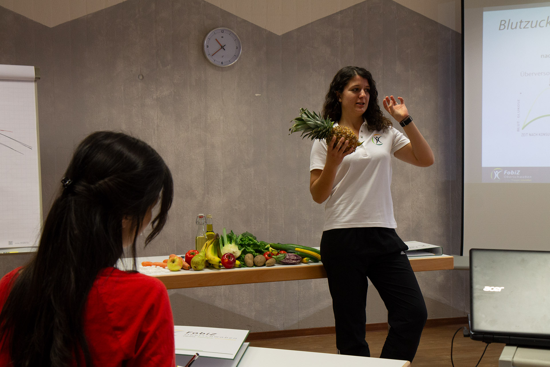 Dozentin hält eine Ananas hoch und spricht vor einer Auslage mit Obst und Gemüse