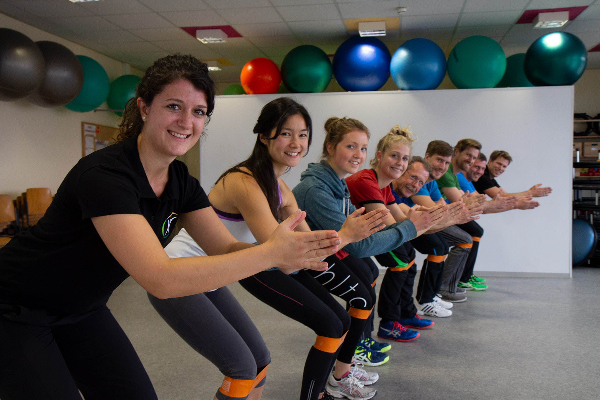 Gruppe macht gemeinsam Kniebeugen mit Fitnessbändern und lächelt in die Kamera